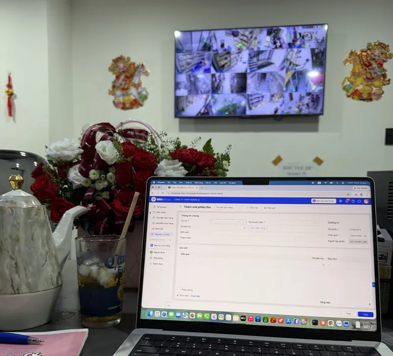 Màn hình camera giám sát nhiều góc kho và máy tính đang mở phần mềm quản lý bán hàng